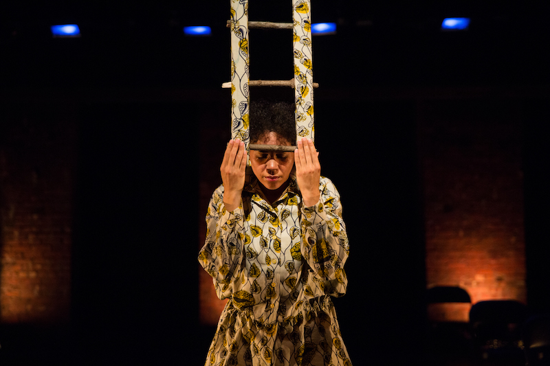 A woman in a yellow printed blouse presses a small prop resembling a mini ladder in front of her. Her head is bowed. The prop is the same pattern as her blouse.